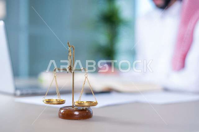 صورة ضبابية لرجل عربي خليجي سعودي يعمل في مجال القضاء، يعمل في قسم المحاماه، امامه ميزان العدل، القانون والمحاماة، يدرس اوراق القضية، استخدام جهاز الحاسوب لانجاز المهام