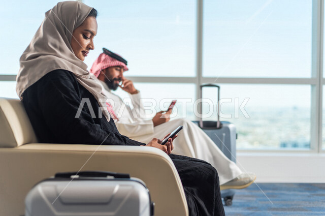 رجل وامراة عربيان خليجيان سعوديان، يستخدمون الهاتف المحمول في تصفح الانترنت، الجلوس في قاعة الانتظار، ايماءات وجه سعيدة، سياحة وسفر