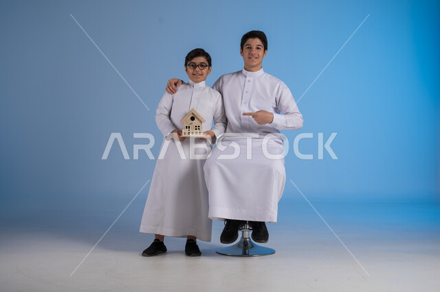 إيماءات السرور والأمل، بيع وشراء العقارات، السعي لامتلاك بيت جديد، بورتريه لشاب عربي خليجي سعودي يرتدي الثوب التقليدي يجلس على كرسي وبجانبه أخيه يحمل بيديه مجسم خشبي صغير لمنزل يشير اليه بأصبعه السبابة، خلفية زرقاء