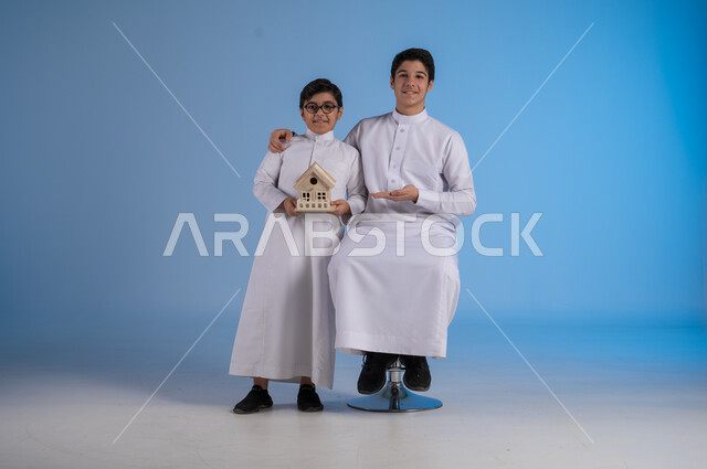السعي لامتلاك بيت جديد، بيع وشراء العقارات، الروابط الأسرية القوية، بورتريه لشاب عربي خليجي سعودي يرتدي الثوب التقليدي يجلس على كرسي وبجانبه اخيه يحمل بيديه مجسم خشبي صغير لمنزل بإيماءات السرور والأمل, خلفية زرقاء