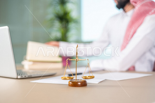 A blurry picture of a Saudi Arabian Gulf man working in the field of justice, wearing traditional Saudi dress, in front of him is the scale of justice, studying case papers, using a computer to complete tasks