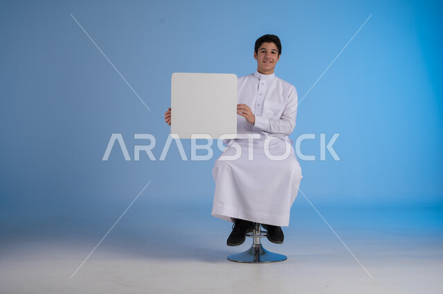 Use of illustrations and educational tools, advertisements and marketing offers, portrait of a young Saudi Arabian Gulf man wearing traditional dress holding a square white board with gestures of pleasure, white square mockup, blank billboard, full-length body image, blue background