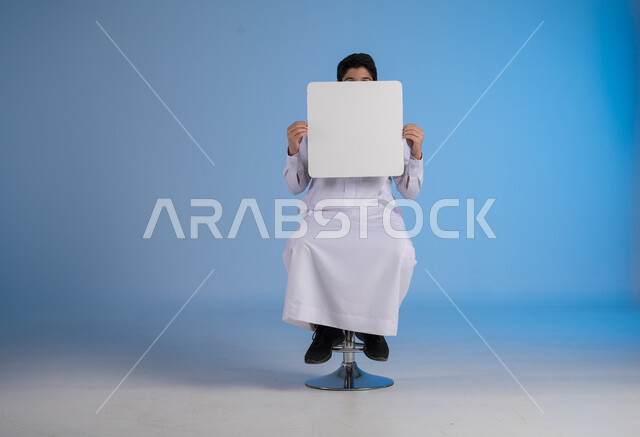 إستخدام لوحات توضيحية ووسائل تعليمية، إعلانات وعروضات تسويقية، بورتريه لشاب عربي سعودي خليجي يرتدي الثوب التقليدي يحمل بيده لوحه بيضاء مربعة يضعها أمام رأسه، موك اب مربع أبيض، لوح إعلاني فارغ،  خلفية زرقاء