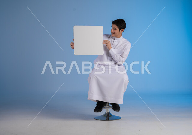 Advertisements and marketing offers, using explanatory panels and educational tools, portrait of a young Saudi Arabian Gulf man wearing traditional dress holding a square white board in his hand and looking at it with gestures of pleasure, white square mockup, blank billboard, blue background