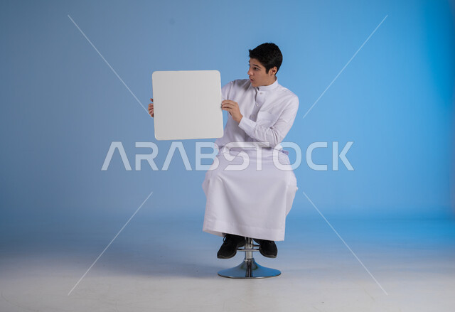 إستخدام لوحات توضيحية ووسائل تعليمية، إعلانات وعروضات تسويقية، بورتريه لشاب عربي سعودي خليجي يرتدي الثوب التقليدي يحمل بيده لوحه بيضاء مربعة ينظر اليها بايماءات السرور، موك اب مربع أبيض، لوح إعلاني فارغ، خلفية زرقاء