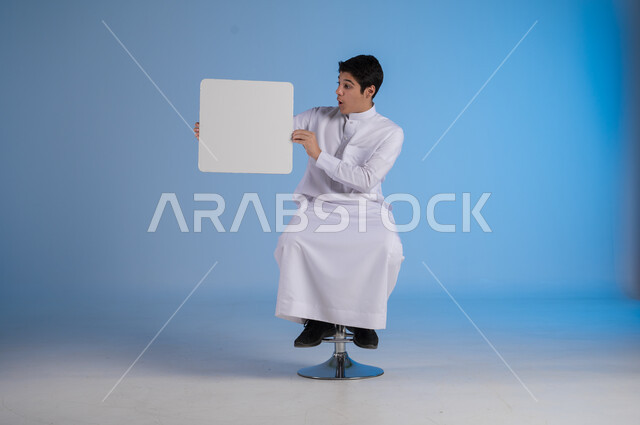 إعلانات وعروضات تسويقية، إستخدام لوحات توضيحية ووسائل تعليمية، بورتريه لشاب عربي سعودي خليجي يرتدي الثوب التقليدي يحمل بيده لوحه بيضاء مربعة ينظر اليها يايماءات السرور، موك اب مربع أبيض، لوح إعلاني فارغ، خلفية زرقاء