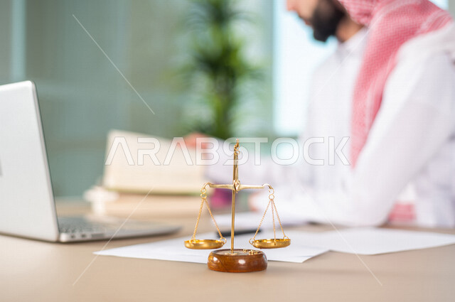 صورة ضبابية لرجل عربي خليجي سعودي يعمل في مجال القضاء، يعمل في قسم المحاماه، امامه ميزان العدل، القانون والمحاماة، يدرس اوراق القضية، استخدام جهاز الحاسوب لانجاز المهام