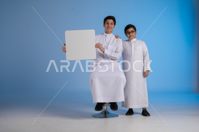 إستخدام لوحات توضيحية ووسائل تعليمية، إعلانات وعروضات تسويقية، بورتريه لشاب عربي سعودي خليجي يرتدي الثوب التقليدي يحمل بيده لوحه بيضاء مربعة وبجانبه أخيه، موك اب مربع أبيض، لوح إعلاني فارغ، صورة الجسم بالطول الكامل, خلفية زرقاء