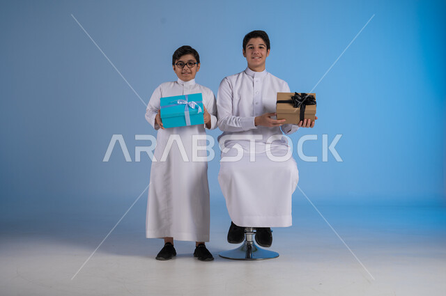 أعياد ومناسبات سعيدة، الشعور بالسرور والفرح, بورتريه لأخوين عربيين سعوديين يرتديان الثوب التقليدي يحملان صندوق هداية مغلف ينظران إلى الكاميرا بتعابير السعادة، تقديم الهدايا والتهنئات, خلفية زرقاء