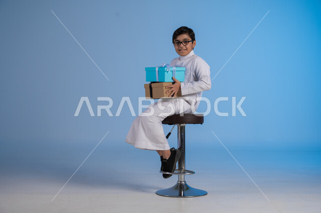 أعياد ومناسبات سعيدة، الشعور بالسعادة والفرح, بورتريه لفتى عربي خليجي سعودي مبتسم يرتدي الثوب التقليدي ينظر إلى الكاميرا يحمل بيديه علب هدايا مغلفة، تقديم الهدايا والتهنئات, خلفية زرقاء