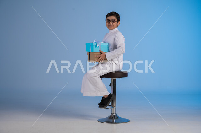 أعياد ومناسبات سعيدة، الشعور بالسعادة والفرح, بورتريه لفتى عربي خليجي سعودي مبتسم يرتدي الثوب التقليدي ينظر إلى الكاميرا يحمل بيديه علب هدايا مغلفة، تقديم الهدايا والتهنئات, خلفية زرقاء