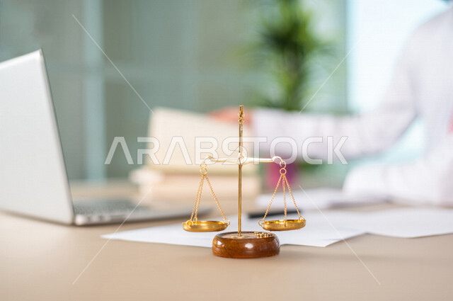A blurry picture of a Saudi Arabian Gulf man working in the field of justice, wearing traditional Saudi dress, in front of him is the scale of justice, studying case papers, using a computer to complete tasks