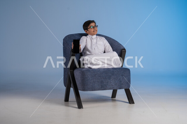 عرض هاتف محمول بشاشة سوداء فارغة، استخدام الأجهزة التقنية الحديثة المتطورة، بورتريه لطفل عربي خليجي سعودي مبتسم يرتدي الثوب التقليدي يجلس على كنبة يمسك بيده الجوال، ايماءات وتعابير السرور والسعادة، خلفية زرقاء