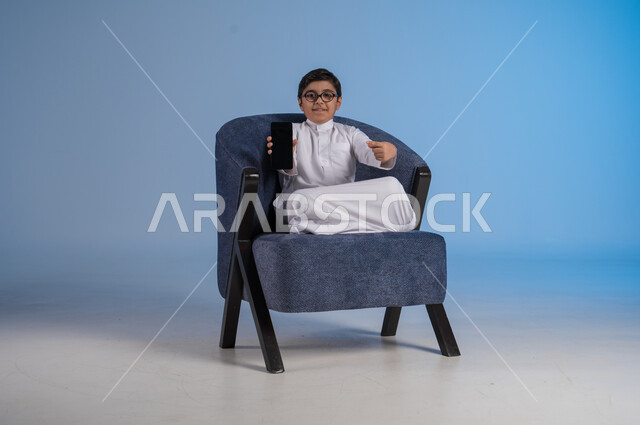 Mobile phone display with blank black screen, using modern advanced technical devices, portrait of a smiling Saudi Arabian Gulf child wearing traditional dress sitting on a sofa holding mobile phone in his hand, gestures and expressions of pleasure and happiness, blue background