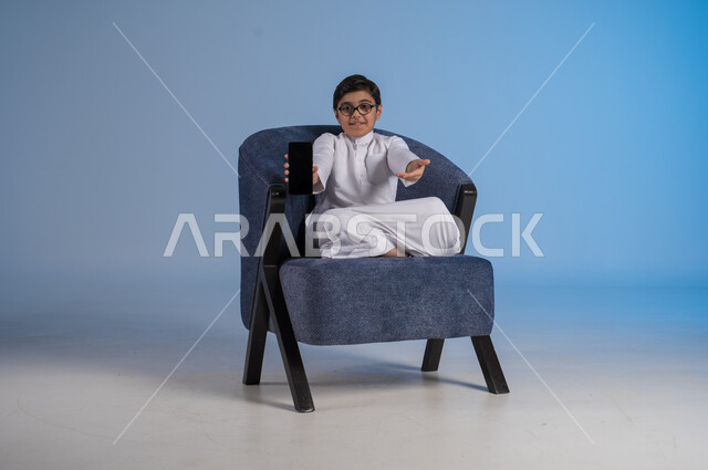 عرض هاتف محمول بشاشة سوداء فارغة، استخدام الأجهزة التقنية الحديثة المتطورة، بورتريه لطفل عربي خليجي سعودي مبتسم يرتدي الثوب التقليدي يجلس على كنبة يمسك بيده الجوال، ايماءات وتعابير السرور والسعادة، خلفية زرقاء