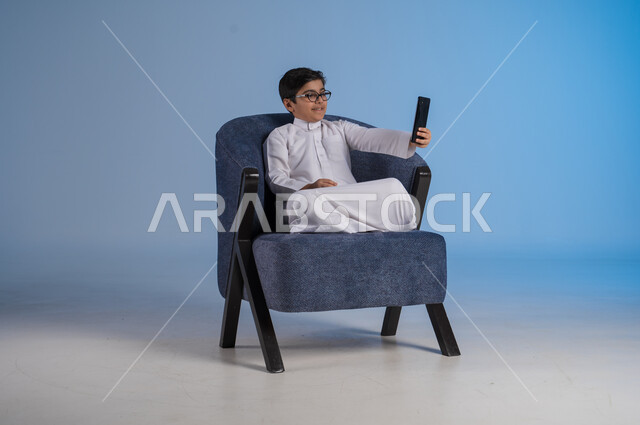 استخدام التكنولوجيا والأجهزة التقنية الحديثة المتطورة، ايماءات السعادة والفرح، بورتريه مقرب لطفل عربي خليجي سعودي مبتسم يرتدي الثوب التقليدي يمسك بيده الهاتف المحمول يجري مكالمة هاتفية، خلفية زرقاء