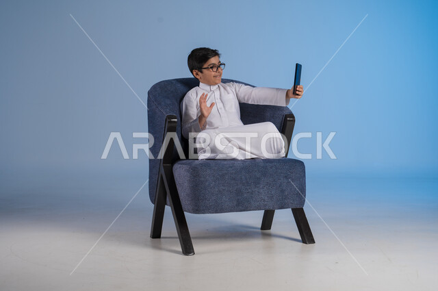 استخدام التكنولوجيا والأجهزة التقنية الحديثة المتطورة، ايماءات السعادة والفرح، بورتريه مقرب لطفل عربي خليجي سعودي مبتسم يرتدي الثوب التقليدي يمسك بيده الهاتف المحمول يجري مكالمة هاتفية، خلفية زرقاء