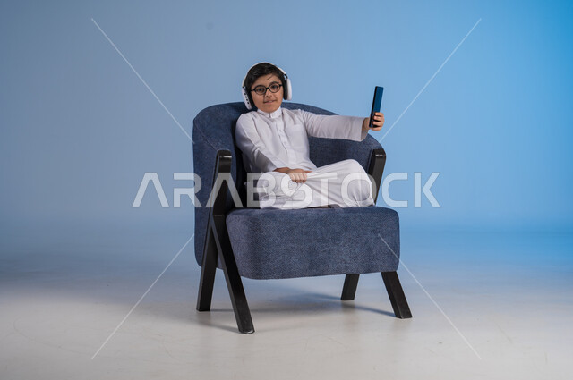 استخدام تقنية الهيدفون الحديثة, قضاء وقت ممتع بالاستماع إلى المقاطع الموسيقية المفضلة, بورتريه لشاب عربي خليجي سعودي يرتدي الثوب التقليدي يجلس على كنبه مريحة يضع سماعة الرأس الصوتية ويمسك بيده الهاتف المحمول, خلفية زرقاء