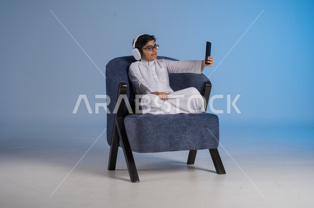 استخدام تقنية الهيدفون الحديثة, قضاء وقت ممتع بالاستماع إلى المقاطع الموسيقية المفضلة, بورتريه لشاب عربي خليجي سعودي يرتدي الثوب التقليدي يجلس على كنبه مريحة يضع سماعة الرأس الصوتية ويمسك بيده الهاتف المحمول, خلفية زرقاء