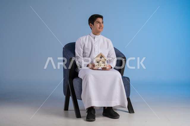 النظر الى شيء ما بإيماءات السرور والسعادة، السعي لامتلاك بيت جديد، بيع وشراء العقارات، بورتريه لشاب عربي خليجي سعودي يرتدي الثوب التقليدي يجلس على كنبة مريحة ويحمل بيديه مجسم خشبي صغير لمنزل، خلفية زرقاء
