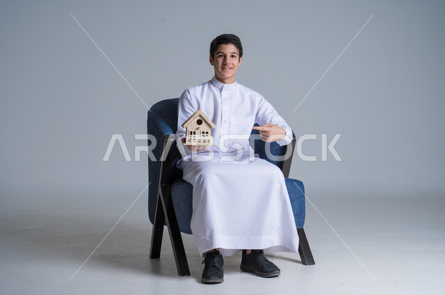السعي لامتلاك بيت جديد، بيع وشراء العقارات، بورتريه لشاب عربي خليجي سعودي يرتدي الثوب التقليدي يجلس على كنبة مريحة ويحمل بيديه مجسم خشبي صغير لمنزل يشير اليه بأصبعه السبابة، إيماءات السرور والسعادة،  خلفية زرقاء