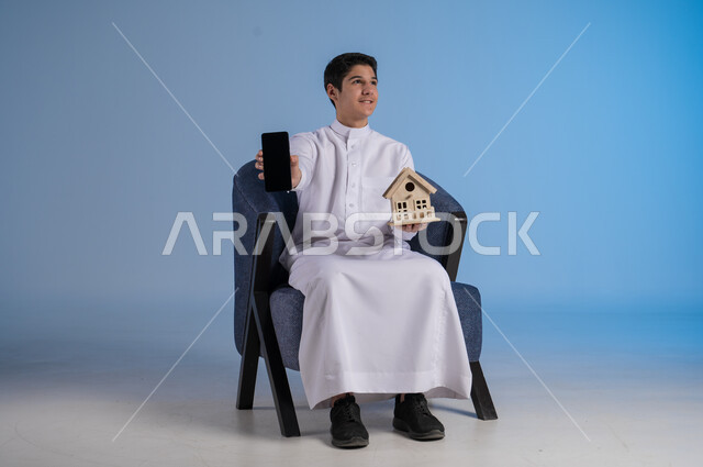 استخدام التقنيات الحيثة المتطورة، السعي لامتلاك بيت جديد، بيع وشراء العقارات، بورتريه لشاب عربي خليجي سعودي يرتدي الثوب التقليدي يجلس على كنبة مريحة ويحمل بيديه مجسم خشبي لمنزل وهاتف بشاشة سوداء، إيماءات السرور والسعادة،  خلفية زرقاء