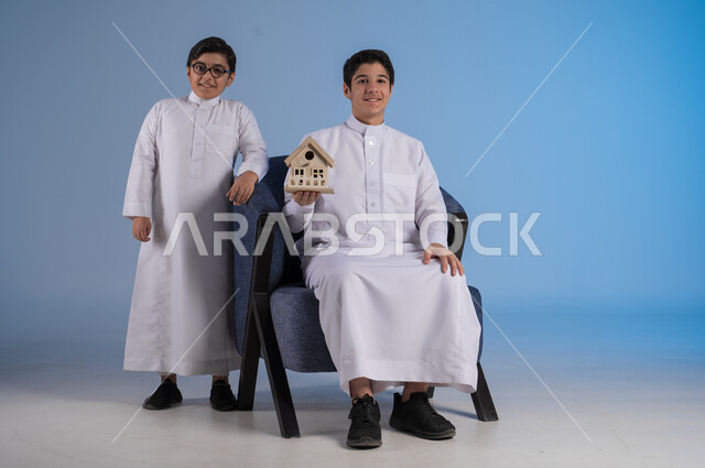 السعي لامتلاك بيت جديد، بيع وشراء العقارات، بورتريه لشاب عربي خليجي سعودي يرتدي الثوب التقليدي يجلس على كنبة مريحة ويحمل بيديه مجسم خشبي صغير لمنزل، إيماءات السرور والسعادة،  خلفية زرقاء