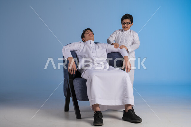 مفهوم المساعدة والتعاون بين الاخوين، ايماءات الراحة والاسترخاء، بورتريه لطفل عربي خليجي سعودي يرتدي الثوب التقليدي يجلس على كنبية ويقف بجانبه اخيه يقوم بتدليك ذراعه، مفهوم المودة والحنان، خلفية زرقاء