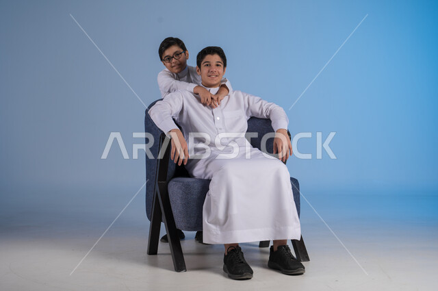 الألفة والمودة بين أفراد السعودية، مفهوم الحب والاحترام بين الاخوين، بورتريه لأخين عربيين خليجيين سعوديين يرتديان الثوب التقليدي ينظران الى الكاميرا بايماءات السعادة والسرور، خلفية زرقاء