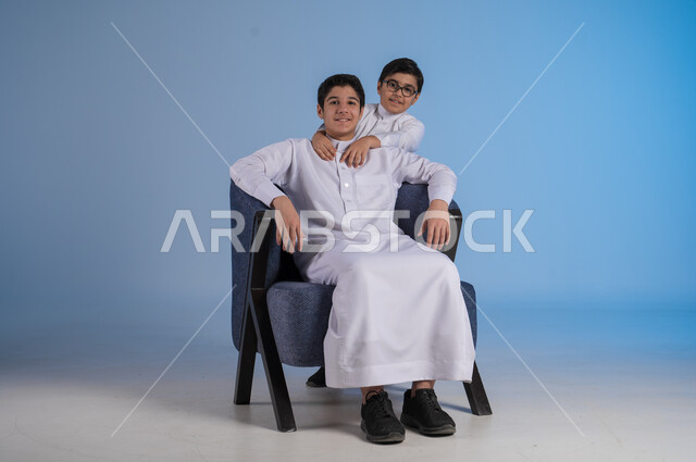 الألفة والمودة بين أفراد السعودية، مفهوم الحب والاحترام بين الاخوين، بورتريه لأخين عربيين خليجيين سعوديين يرتديان الثوب التقليدي ينظران الى الكاميرا بايماءات السعادة والسرور، خلفية زرقاء