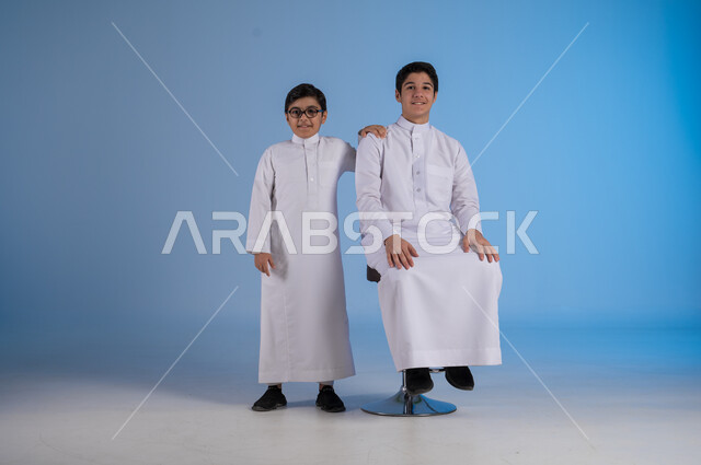 الألفة والمودة بين أفراد السعودية، مفهوم الحب والاحترام بين الاخوين، بورتريه لطفلين عربيين خليجيين سعوديين يرتديان الثوب التقليدي يقفان بجانب بعضهما ينظران الى الكاميرا بايماءات السعادة والسرور، خلفية زرقاء