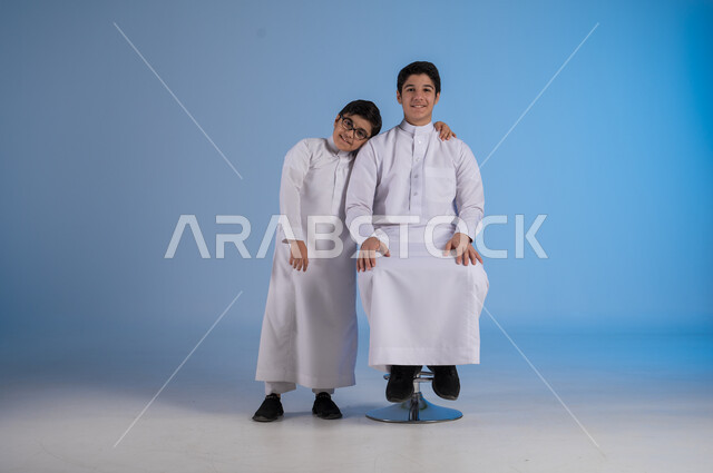 Spreading love and affection between brothers, portrait of a Saudi Arabian Gulf boy wearing traditional dress leaning on his older brother's shoulder, gestures of happiness and pleasure, blue background