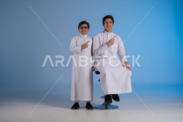 الاشارة باصبع السبابة الى شيء ما، النظر إلى الكاميرا بتعابير السرور، بورتريه لأخين عربيان خليجيان سعوديان مبتسمان يرتديان الزي التقليدي يقومان بحركات يد، خلفية زرقاء