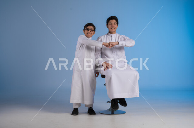 الاتفاق على أمر معين، النظر إلى الكاميرا بتعابير السرور، القبول والرضا بالنتيجة، ايماءات الاعجاب والموافقة، بورتريه لطفلين عربيان خليجيان سعوديان مبتسمان يرتديان الزي التقليدي يقومان بحركات يد تدل على علامة الجودة والامتياز، خلفية زرقاء