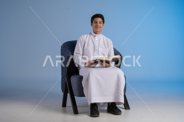 قضاء الوقت بالقراءة والتعلم والثقافة، التمعن والتركيز في الرواية، بورتريه لشاب عربي سعودي خليجي يرتدي الزي السعودي التقليدي يجلس على كرسي يحمل بيده كتاب، خلفية زرقاء