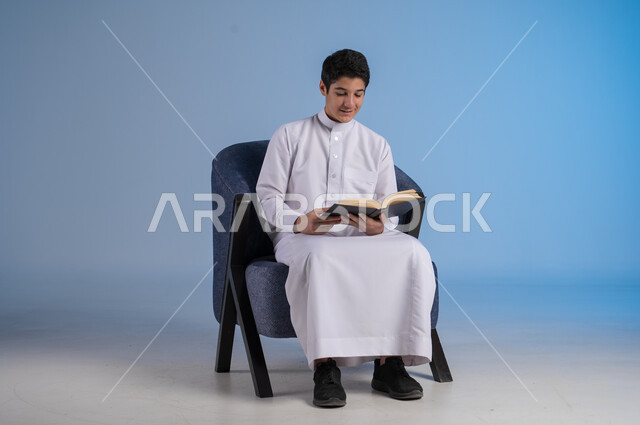 قضاء الوقت بالقراءة والتعلم والثقافة، التمعن والتركيز في الرواية، بورتريه لشاب عربي سعودي خليجي يرتدي الزي السعودي التقليدي يجلس على كرسي يحمل بيده كتاب، خلفية زرقاء