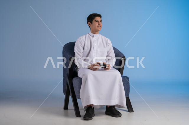 قضاء وقت ممتع ومسلي، التفاعل والاندماج مع ألعاب البلايستيشن، تقنيات حديثة مسلية, بورتريه لشاب عربي سعودي خليجي يرتدي الزي التقليدي يجلس على كرسي مريح ويحمل بيده عصا التحكم  ويلعب بألعاب الفيديو ، خلفية زرقاء