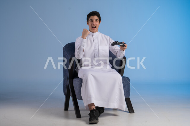 النظر الى الكاميرا بايماءات السرور، قضاء وقت ممتع ومسلي، التفاعل والاندماج مع ألعاب البلايستيشن، تقنيات حديثة مسلية, بورتريه لشاب عربي سعودي خليجي يرتدي الزي التقليدي يجلس على كرسي مريح ويحمل بيده عصا التحكم  ويلعب بألعاب الفيديو ، خلفية زرقاء