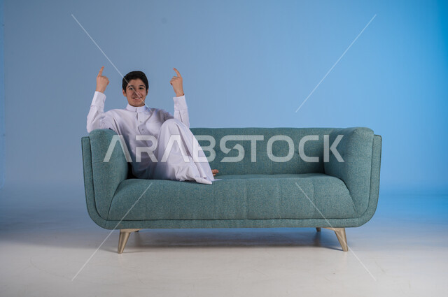 الاشارة الى شيء ما في الأعلى, النظر الى الكاميرا بايماءات السعادة، بورتريه لفتى عربي خليجي سعودي يرتدي الثوب التقليدي يرفع أصابع يده بإيماءات السرور, صورة الجسم بالطول الكامل، خلفية زرقاء