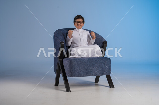 Concept of winning and triumph, portrait of a Saudi Arabian Gulf boy wearing traditional dress and glasses raising his hands with gestures of happiness and pleasure, joy for achieving something, blue background