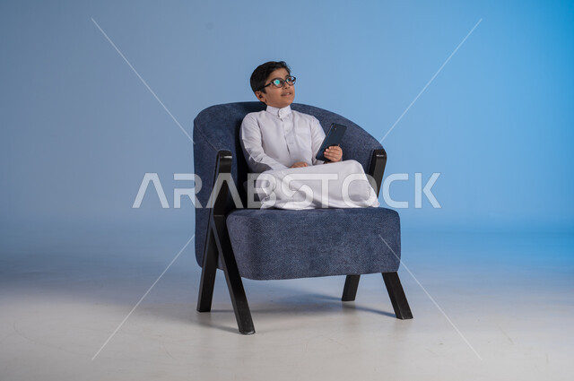 التواصل مع الاصدقاء عبر الهاتف المحمول، استخدام التقنيات الحديثة والتكنولوجيا، بورتريه لشاب عربي سعودي خليجي يرتدي الثوب التقليدي يحمل بيده الموبايل يجلس على كرسي مريح ينظر يحاول النظر الى شيء ما بالأعلى، صورة الجسم بالطول الكامل خلفية زرقاء