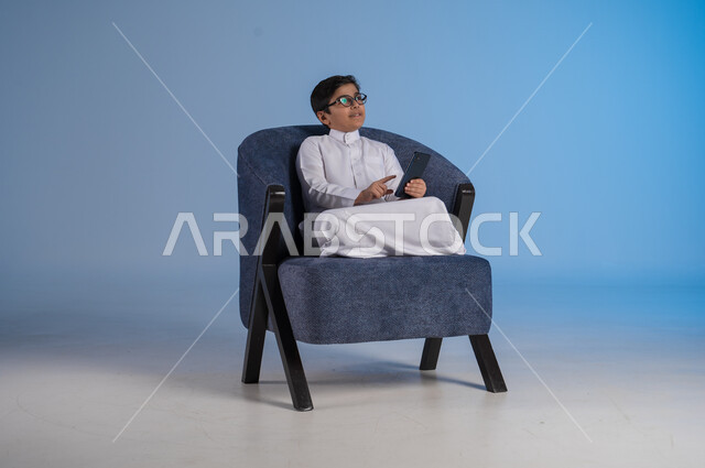 Communicating with friends via mobile phone, using modern techniques and technology, portrait of a Saudi Arabian Gulf boy wearing traditional dress holding a mobile phone in his hand sitting on a comfortable chair looking trying to look at something above, blue background