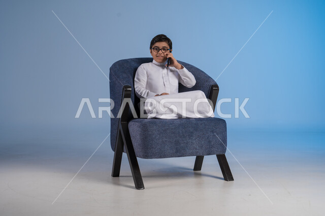 التواصل مع الاصدقاء عبر الهاتف المحمول، استخدام التقنيات الحديثة والتكنولوجيا، بورتريه لفتى عربي سعودي خليجي يرتدي الثوب التقليدي يحمل بيده الموبايل يجلس على كرسي مريح، خلفية زرقاء