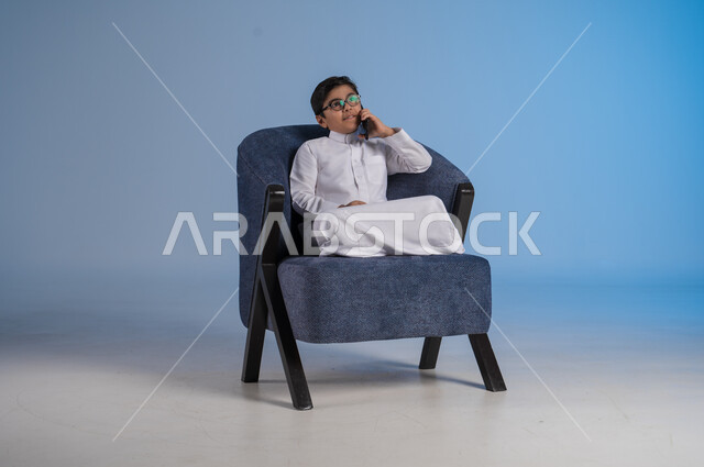 التواصل مع الاصدقاء عبر الهاتف المحمول، استخدام التقنيات الحديثة والتكنولوجيا، بورتريه لفتى عربي سعودي خليجي يرتدي الثوب التقليدي يحمل بيده الموبايل يجلس على كرسي مريح، خلفية زرقاء