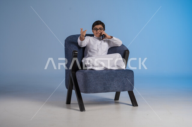 التواصل مع الاصدقاء عبر الهاتف المحمول، استخدام التقنيات الحديثة والتكنولوجيا، بورتريه لفتى عربي سعودي خليجي يرتدي الثوب التقليدي يحمل بيده الموبايل يجلس على كرسي مريح، خلفية زرقاء