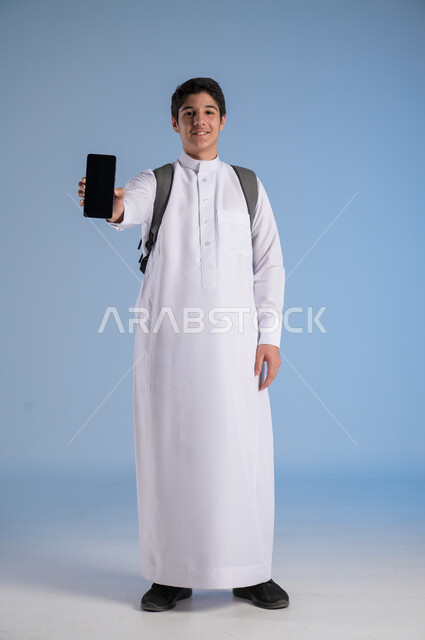 استخدام التكنولوجيا الحديثة للتسوق الالكتروني عبر الانترنت, عرض تطبيقات وخدمات الهاتف المحمول, بورتريه لشاب عربي خليجي سعودي مبتسم يرتدي الثوب التقليدي يحمل بيده جوال بشاشه سوداء فارغة, النظر إلى الكاميرا بإيماءات السرور, صورة الجسم بالطول الكامل، خلفيه زرقاء