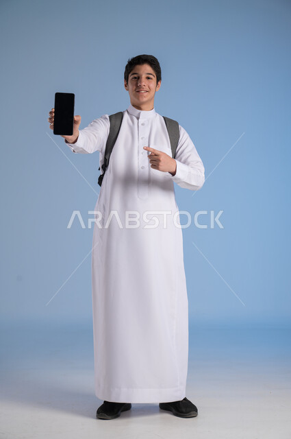 استخدام التكنولوجيا الحديثة للتسوق الالكتروني عبر الانترنت, عرض تطبيقات وخدمات الهاتف المحمول, بورتريه لشاب عربي خليجي سعودي مبتسم يرتدي الثوب التقليدي يحمل بيده جوال بشاشه سوداء فارغة ويشير إليه, النظر إلى الكاميرا بإيماءات السرور, صورة الجسم بالطول الكامل، خلفيه زرقاء