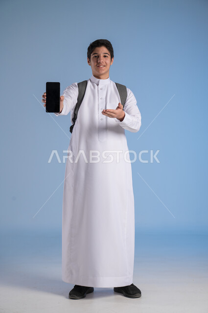 استخدام التكنولوجيا الحديثة للتسوق الالكتروني عبر الانترنت, عرض تطبيقات وخدمات الهاتف المحمول, بورتريه لشاب عربي خليجي سعودي مبتسم يرتدي الثوب التقليدي يحمل بيده جوال بشاشه سوداء فارغة ويشير إليه, النظر إلى الكاميرا بإيماءات السرور, صورة الجسم بالطول الكامل، خلفيه زرقاء