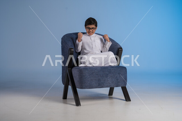 الغضب من شيء ما, فتى سعودي يجلس على كرسي بايماءات يدين ووجه تدل على الضجر و الاستياء وعدم الرضا, بورتريه لطفل خليجي سعودي يرتدي الثوب التقليدي والنظارات الطبية يرفع يده للاعلى بتعابير العصبية, صورة الجسم بالطول الكامل، خلفية زرقاء