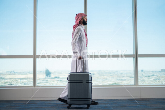 A Saudi Arabian Gulf businessman holding the travel bag and towing it, preparing to travel on a business trip, waiting lounge at the airport, tourism and travel, spending fun times